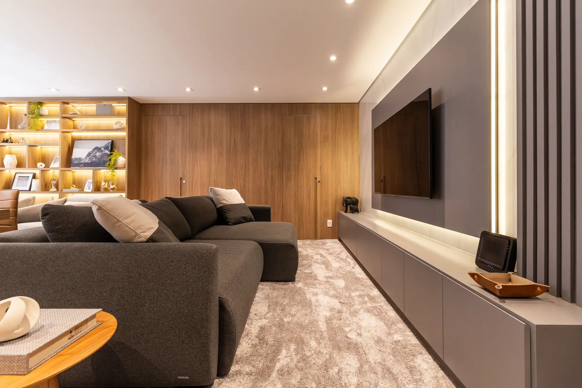 Sala de TV com estante iluminada, painel de madeira com portas, painel da TV, rack e ripado executados pela FeP